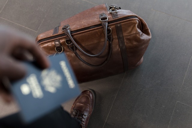 Imagem de homem com passaporte na mão e mala de viagem no chão.